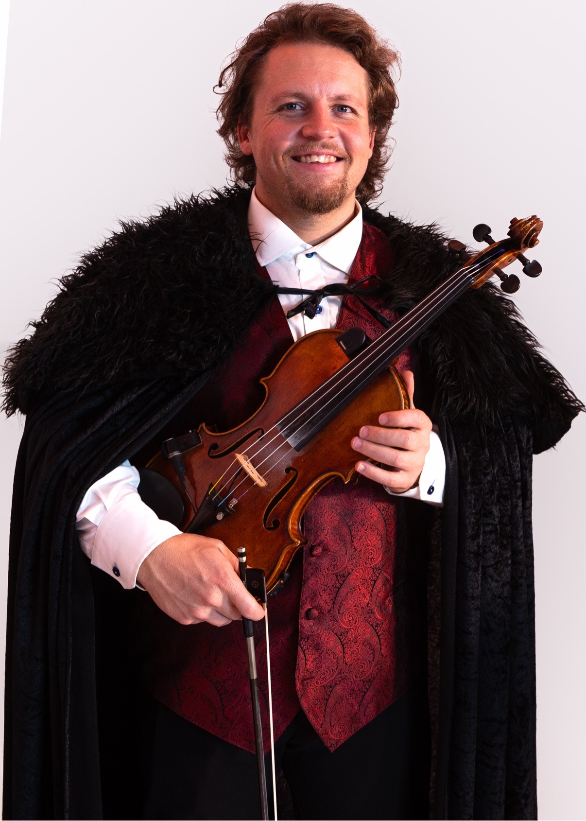 Jake smiling in a red paisley vest and black cape while holding his violin