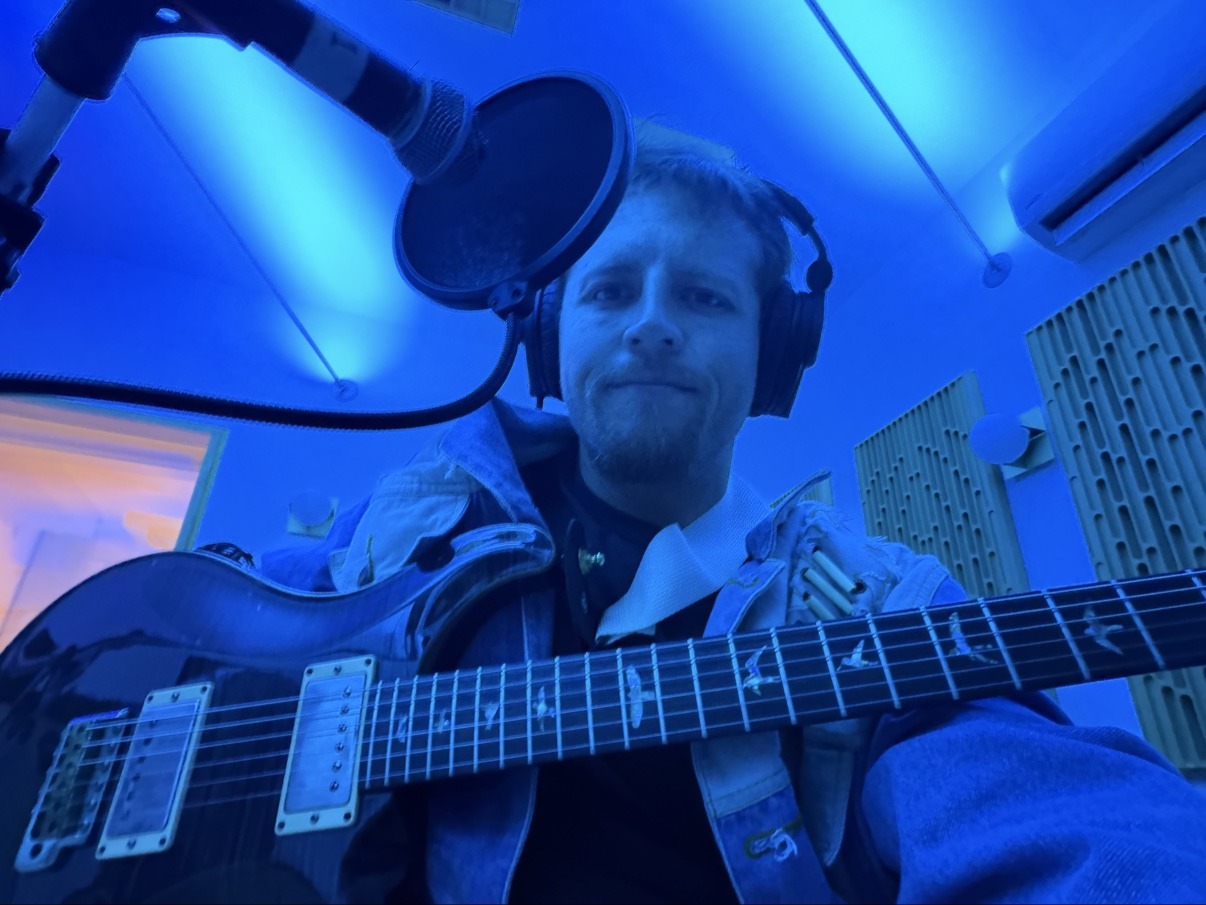 Jake with a guitar and headphones at a blue-lit studio microphone
