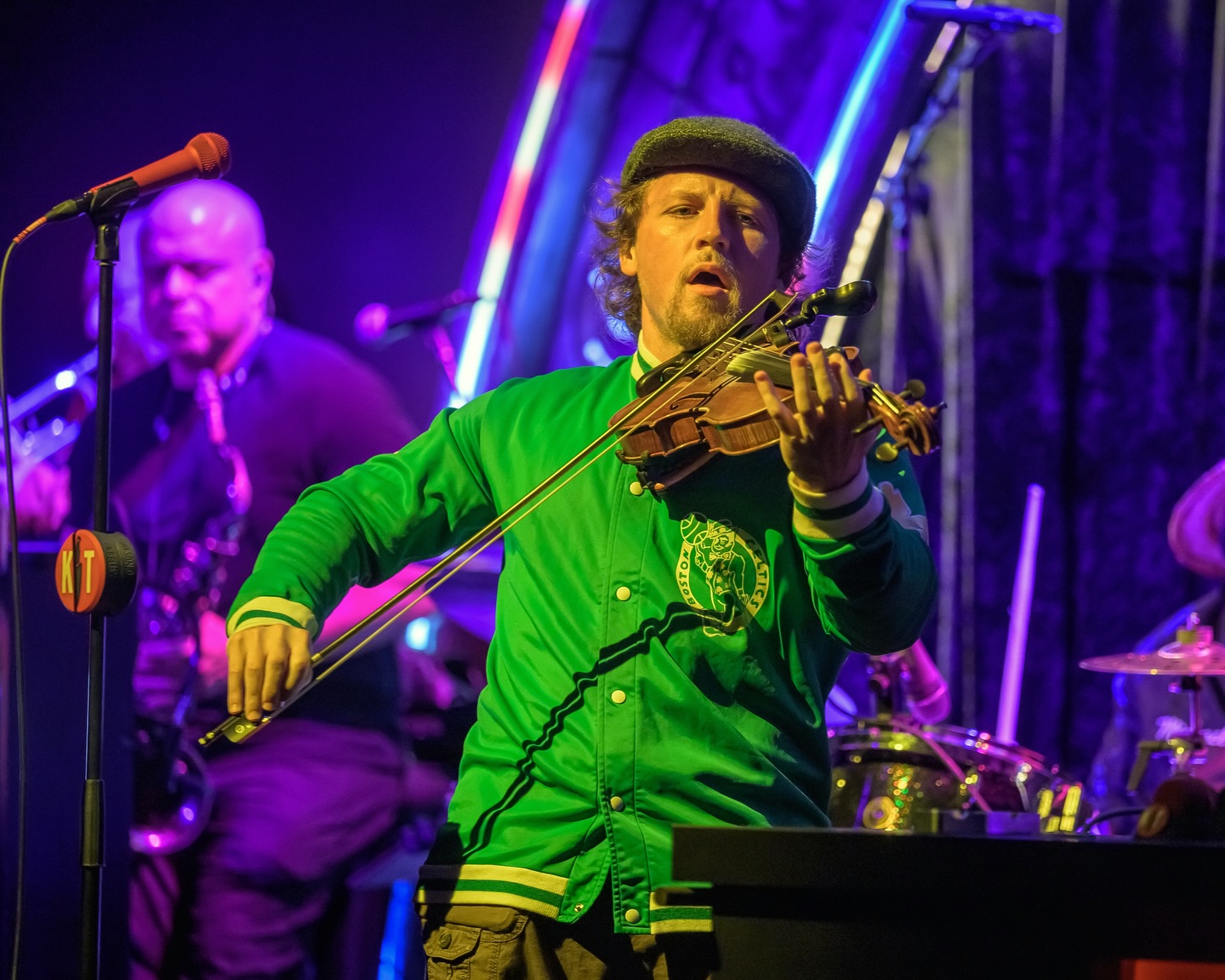 Jake Cross playing violin on the Kill Tony stage in a green jacket